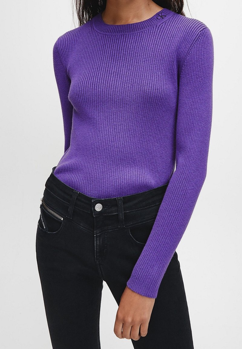 Suéter morado acanalado con mangas largas y cuello redondo. Combinado con unos jeans de mezclilla negros que presentan un detalle de bolsillo con cremallera.