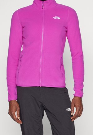 Osoba ubrana w jasnoróżową polarową kurtkę rozpinaną na zamek oraz czarne spodnie, obie z logo The North Face na piersi i udzie.