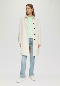 Jasno beżowy trencz o prostym kroju, z guzikami z przodu i kieszeniami bocznymi, nałożony na miętowy sweter i niebieskie dżinsy. Białe trampki.