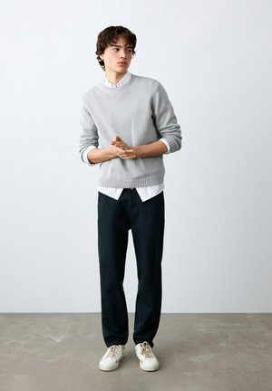 Giovane uomo con maglione grigio chiaro sopra una camicia bianca, pantaloni neri e scarpe da ginnastica bianche, in piedi contro un muro bianco semplice.