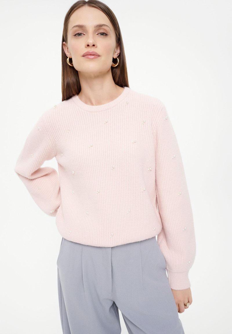 Hellrosa Rippstrickpullover mit Perlenakzenten, rundem Ausschnitt und langen Ärmeln. Weicher Stoff, kombiniert mit grauen Hosen.