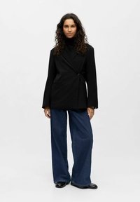 Juodas pritaikytas švarkas su vienu mygtuku, pasižymintis iškarpytomis apykaklėmis ir priekinių kišenėmis, derinamas su plačiais mėlynos džinsinio audinio keliais.