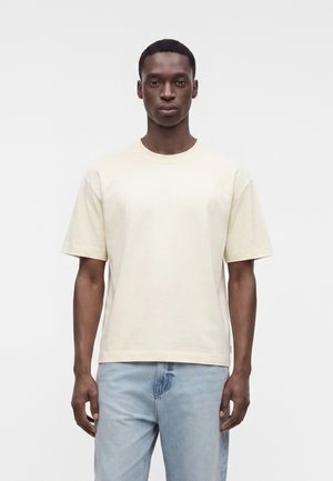 Homme debout devant un fond uni, portant un t-shirt à manches courtes beige clair et un jean bleu clair, faisant face avec une expression neutre.