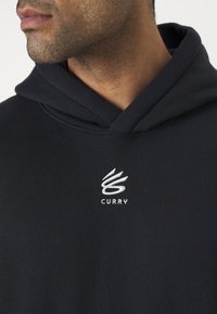 Svart hettegenser laget av mykt materiale, med et hvitt frontlogo, avslappet passform og snor i hetten.
