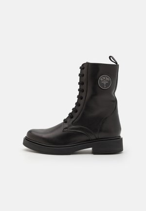Stivale militare in pelle nera con punta tonda, chiusura con lacci e un patch con logo circolare su un lato. Presenta una suola spessa e una texture liscia.