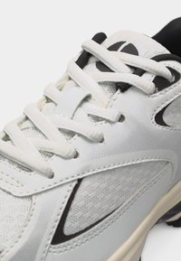 Chaussure de sport blanche et noire avec des panneaux en maille texturée, des lacets blancs passés dans des œillets, et un col de cheville noir rembourré.