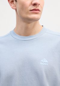 Svetlo modrá mikina vyrobená z jemnej bavlny, s rebrovaným krkom a malým bielym logom hory so slovom "BOSS" na hrudi.