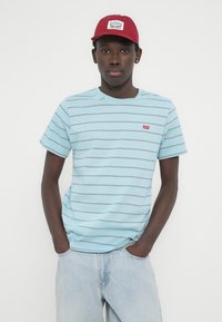 Γαλάζιο ριγέ t-shirt με κοντά μανίκια και στρογγυλή λαιμόκοψη, συνδυασμένο με κόκκινο καπέλο που φέρει λευκό λογότυπο σε μπαλώμα. Τζιν παντελόνι με άνετη εφαρμογή.