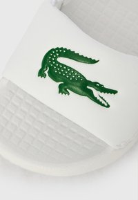 Λευκό σανδάλι slide με υφή στη σόλα, που διαθέτει πράσινο λογότυπο κροκόδειλου σε λείο λουράκι. Καθαρός σχεδιασμός, μινιμαλιστική εμφάνιση.