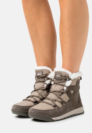 Nogi w beżowo-brązowych zimowych butach z białym futerkiem w środku i sznurowadłami w kształcie krzyżyków, stojące na gładkim białym tle.