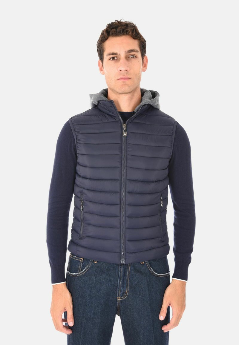 Uomo che indossa un gilet trapuntato blu navy con zip frontale e cappuccio grigio, un maglione blu navy e jeans blu, in piedi su uno sfondo bianco.
