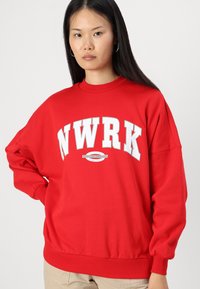 Felpa rossa con scollo a giro, vestibilità oversized e maniche lunghe. Presenta la scritta bianca "NWRK" e un logo grafico stampato sul davanti.