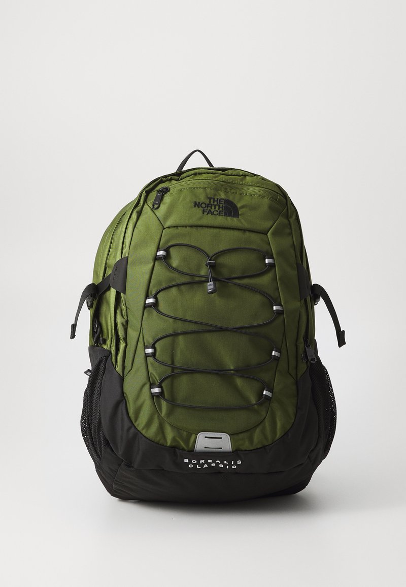 Mochila verde com base preta, apresentando uma superfície texturizada, sistema de corda elástica frontal, compartimentos com zíper e bolsos laterais em malha.