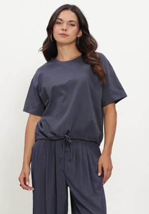 Vrouw met lang donker haar, die een marineblauw shirt met korte mouwen en geplooide broek draagt, staand tegen een effen lichte achtergrond.