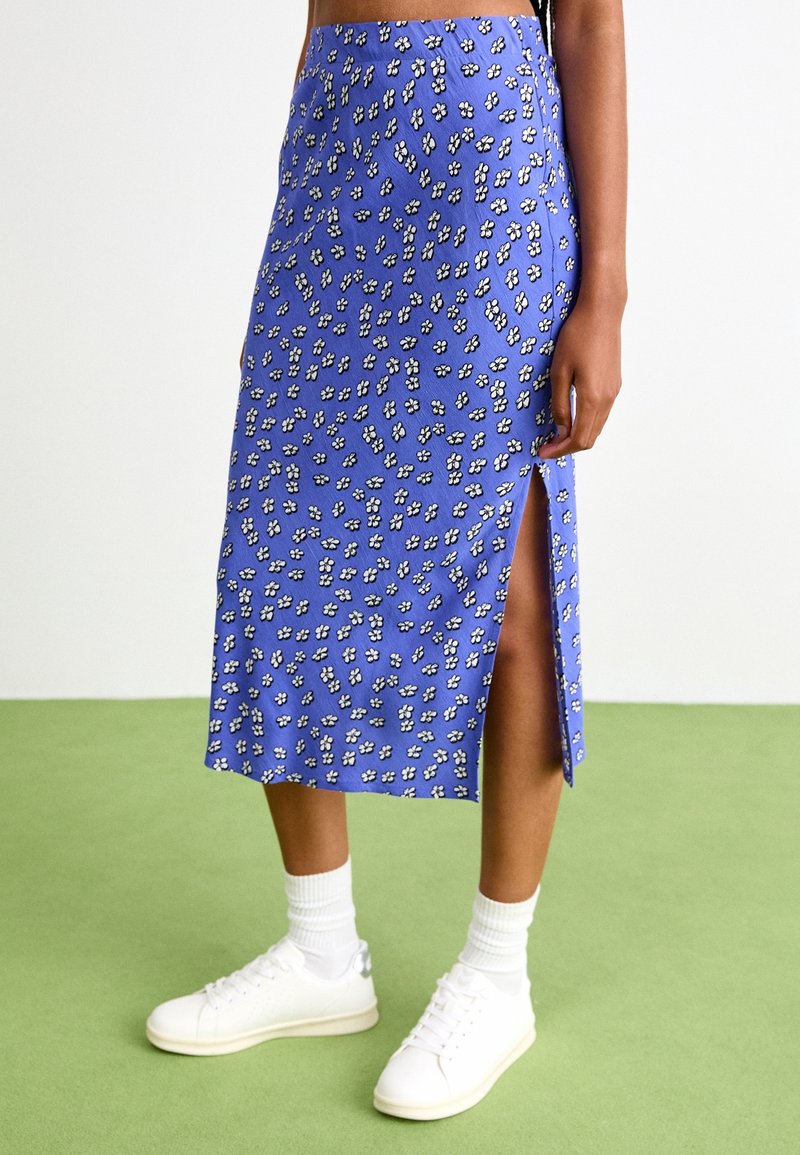 Jupe midi bleu à motif floral avec une fente latérale, en tissu léger. Stylée avec des baskets blanches et des chaussettes côtelées.