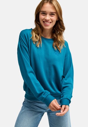 Jonge vrouw glimlacht, draagt een blauwgroene sweatshirt met lange mouwen en lichtblauwe spijkerbroek tegen een effen witte achtergrond.