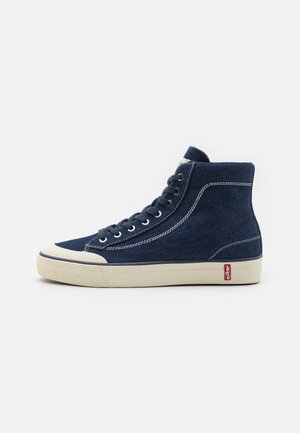 Wysokie granatowe sneakersy z dżinsu z białymi przeszyciami, kremową podeszwą, sznurowadłami, osłoną na palce oraz czerwoną metką Levi's w pobliżu pięty.