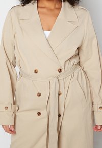 Beżowy trencz z podwójnym zapięciem, dużymi brązowymi guzikami, paskiem w talii i podwiniętymi rękawami. Gładki, teksturowany materiał.
