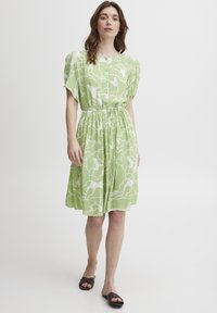Rochie A-line cu mânecă scurtă din material verde deschis cu model abstract alb, talie strânsă și detaliu cu nasture la decolteu.