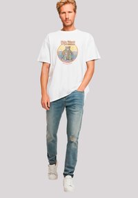 Weißes Baumwoll-T-Shirt mit einem bunten Bären-Graphic und Text; kombiniert mit hellblauen, ausgeblühten Jeans und weißen Turnschuhen.