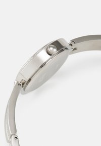 Silverfärgad metallklocka med rund klocka, texturerad bezel och en slät rem. Har en krona med rund topp för justering.