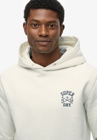 Jasno beżowa bluza z kapturem wykonana z miękkiego materiału, z dużą kieszenią z przodu, kapturem ze sznurkiem oraz naszywanym granatowym logo na klatce piersiowej.