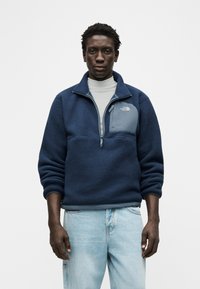 Άνδρας που φοράει μπλε σκούρο φλις μπουφάν με τσέπη στο στήθος και ανοιχτό μπλε τζιν, όρθιος μπροστά σε απλό λευκό φόντο, κοιτάζοντας προς τα εμπρός.