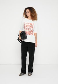 Tricou alb supradimensionat cu design grafic roșu, pantaloni negri cu picioare largi și adidași gri. Geantă neagră ținută într-o mână.