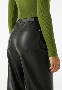 Pantaloni in finta pelle nera con vita alta, tasche posteriori e una texture liscia. Abbinati a un maglione verde a coste aderente.