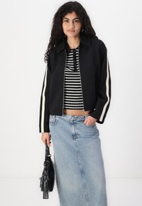 Veste zippée noire avec des rayures crème, portée par-dessus un haut rayé noir et blanc. Associée à une jupe en denim bleu clair et un sac à main noir.