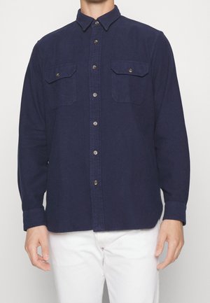 Homme portant une chemise bleu marine boutonnée avec deux poches poitrine et un pantalon blanc, debout devant un fond uni.