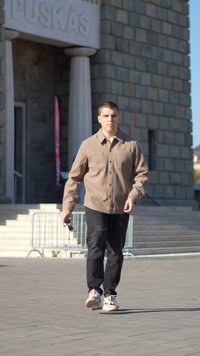 Homme marchant sur une aire pavée près de l'entrée d'un bâtiment en pierre, portant une veste beige, un jean noir déchiré, et tenant des lunettes de soleil dans la main droite.