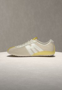 Baskets beige et blanches avec des accents jaunes, lacets à l'avant et semelle crantée, montrées de profil sur une surface et un fond neutres.