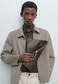 Brun læder clutch med en skrå lynlås, holdt mod en beige uldblandingjakke og en grå skjorte. Glat tekstur, minimalistisk design.