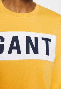 Geel sweatshirt met een witte horizontale streep waarop "GANT" in marineblauw is gedrukt. Zachte textuur, ronde hals en ribgebreide manchetten.