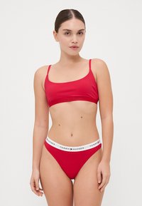Raudonas bikini komplektas, kuris susideda iš apvalaus kaklo liemenėlės ir atitinkamų kelnaičių. Elastinga juosta su „TOMMY HILFIGER“ logotipu. Lygus audinys.