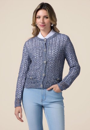 Cardigan en tricot bleu avec fermeture à boutons, motif texturé et deux poches avant. Porté sur une chemise blanche à col et un jean bleu clair.