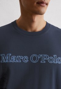 Navy-blaue Baumwoll-T-Shirt mit dem Markennamen "Marc O'Polo" in einem strukturierten hellblauen Druck. Klassischer Rundhalsausschnitt und kurze Ärmel.