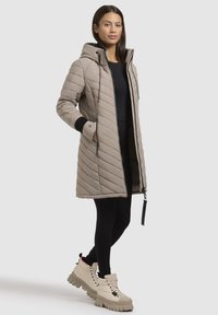 Parka acolchada beige con capucha, de longitud larga, dos bolsillos frontales y puños negros. Usada sobre leggings negros y botines beiges gruesos.