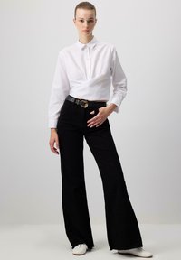 Camicia bianca con bottoni e design avvolgente, abbinata a pantaloni neri a vita alta e svasati. Presenta una cintura con fibbia dorata e scarpe bianche.