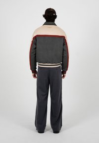 Homme portant une veste noire et beige avec des rayures rouges, un pantalon gris foncé et une casquette noire, debout face à un fond blanc uni.