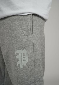 Gray sweatpants feature a textured fabric, side pockets, and a prominent light gray embroidered logo on the left thigh.