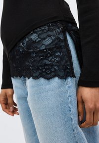 Μαύρη δαντέλα που ξεπροβάλλει από κάτω από μαύρο τοπ πάνω από ανοιχτόχρωμο μπλε τζιν, με ένα χέρι που ακουμπά στο πλάι.