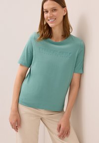 Mujer sonriente con una camiseta de manga corta color teal con el texto en relieve "CIELOS ETERNOS" y pantalones beige, apoyada contra una pared clara.