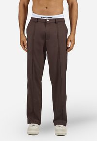 Muškarac u tamnosmešavim krojenim hlačama, belom brendiranim donjem vešu i belim patikama, stoji s opuštenim rukama.