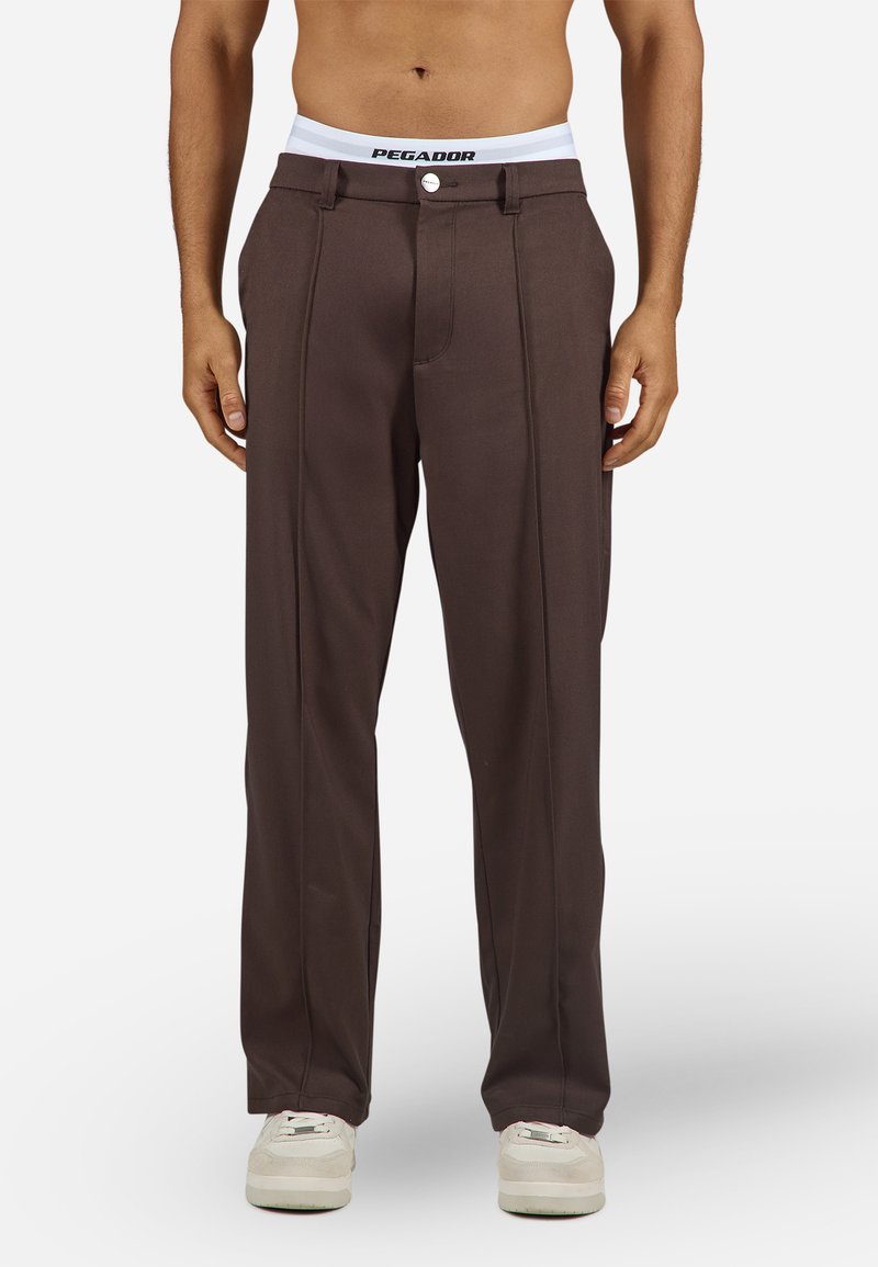 Muškarac u tamnosmešavim krojenim hlačama, belom brendiranim donjem vešu i belim patikama, stoji s opuštenim rukama.