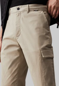 Bež pamukove cargo hlače sa ravnim nogavicama, bočnim džepom i etiketom Calvin Klein na pojasu. Glatka tekstura, minimalistički dizajn.