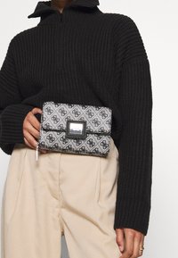Bolso de mano monocromático con un patrón geométrico en negro y plata, con solapa frontal, acentos metálicos y un detalle de cremallera.