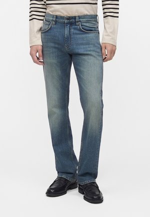 Mand iført blå straight-fit jeans, sorte lædersko og en beige langærmet skjorte med sorte vandrette striber på ærmerne.