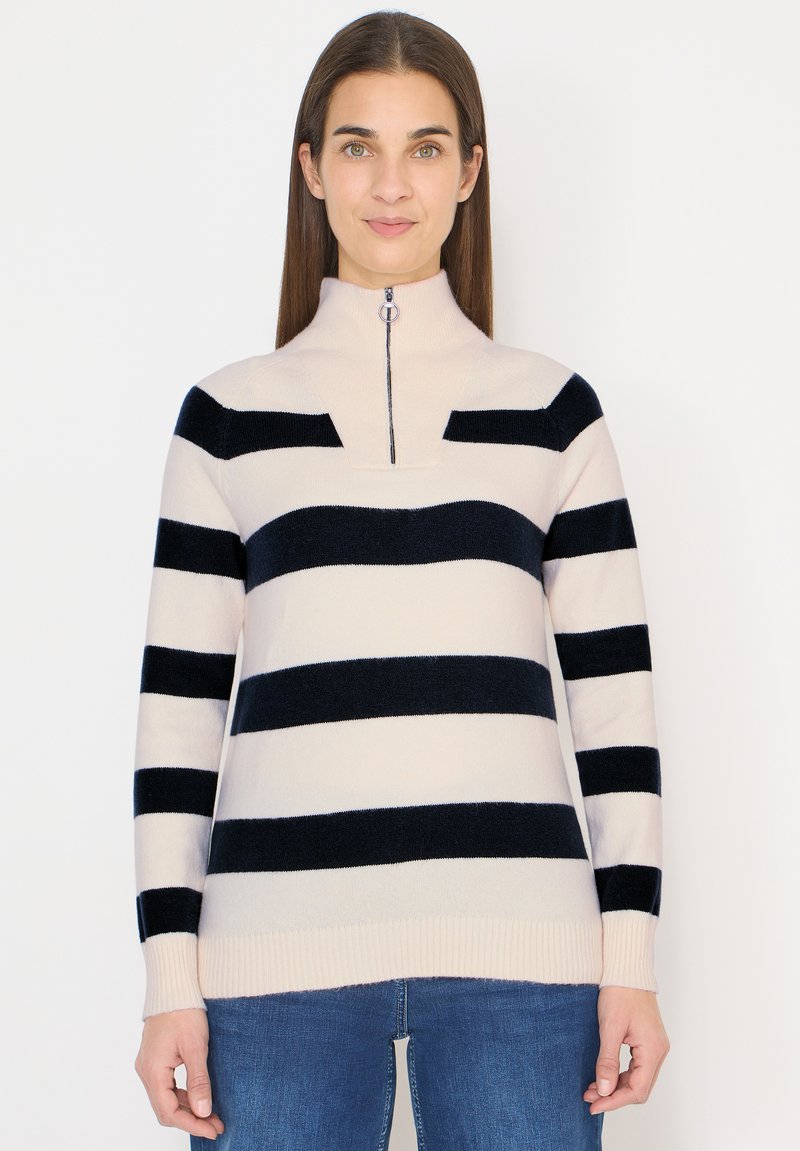 Femme portant un pull clair à rayures horizontales noires avec un col à fermeture éclair partielle, associée à un jean bleu, debout devant un fond blanc.